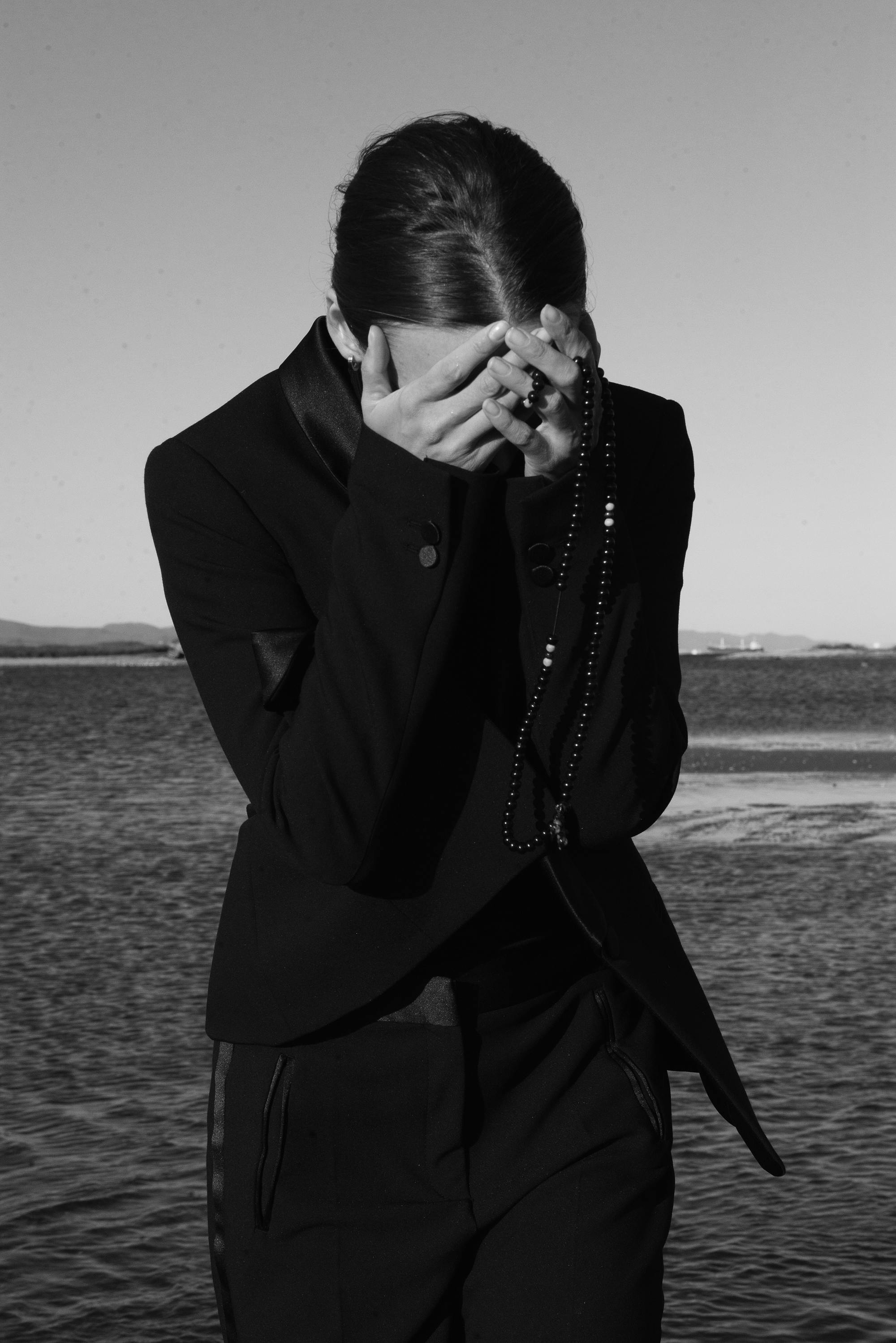 Black and white portrait of a person in elegant fashion, covering face with hands holding beads.