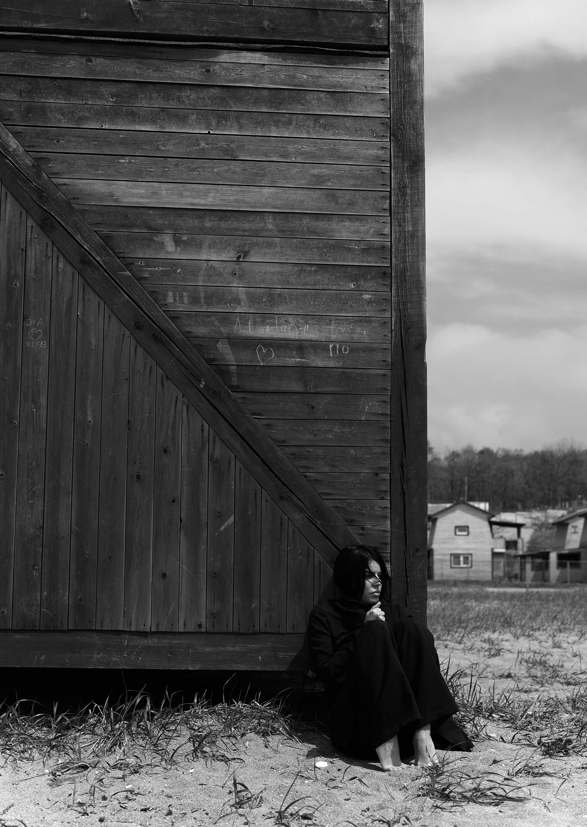 A contemplative woman in monochrome sits beside a wooden building, exuding a moody atmosphere.