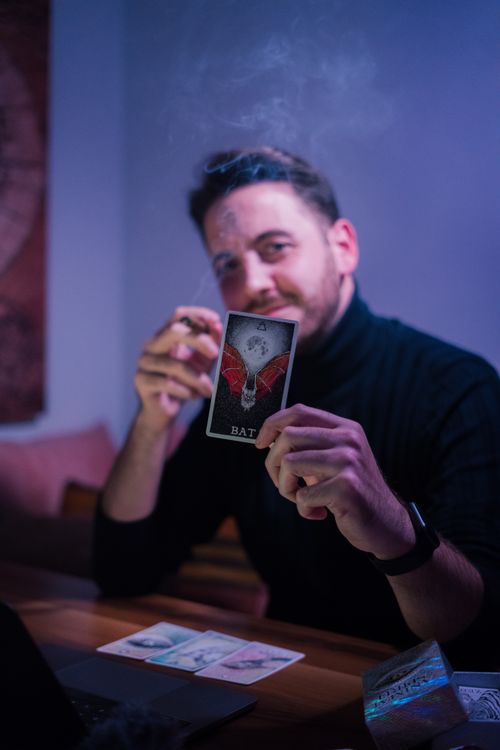 Free Male fortune teller showing tarot card and looking at camera while sitting at table in semi dark room Stock Photo