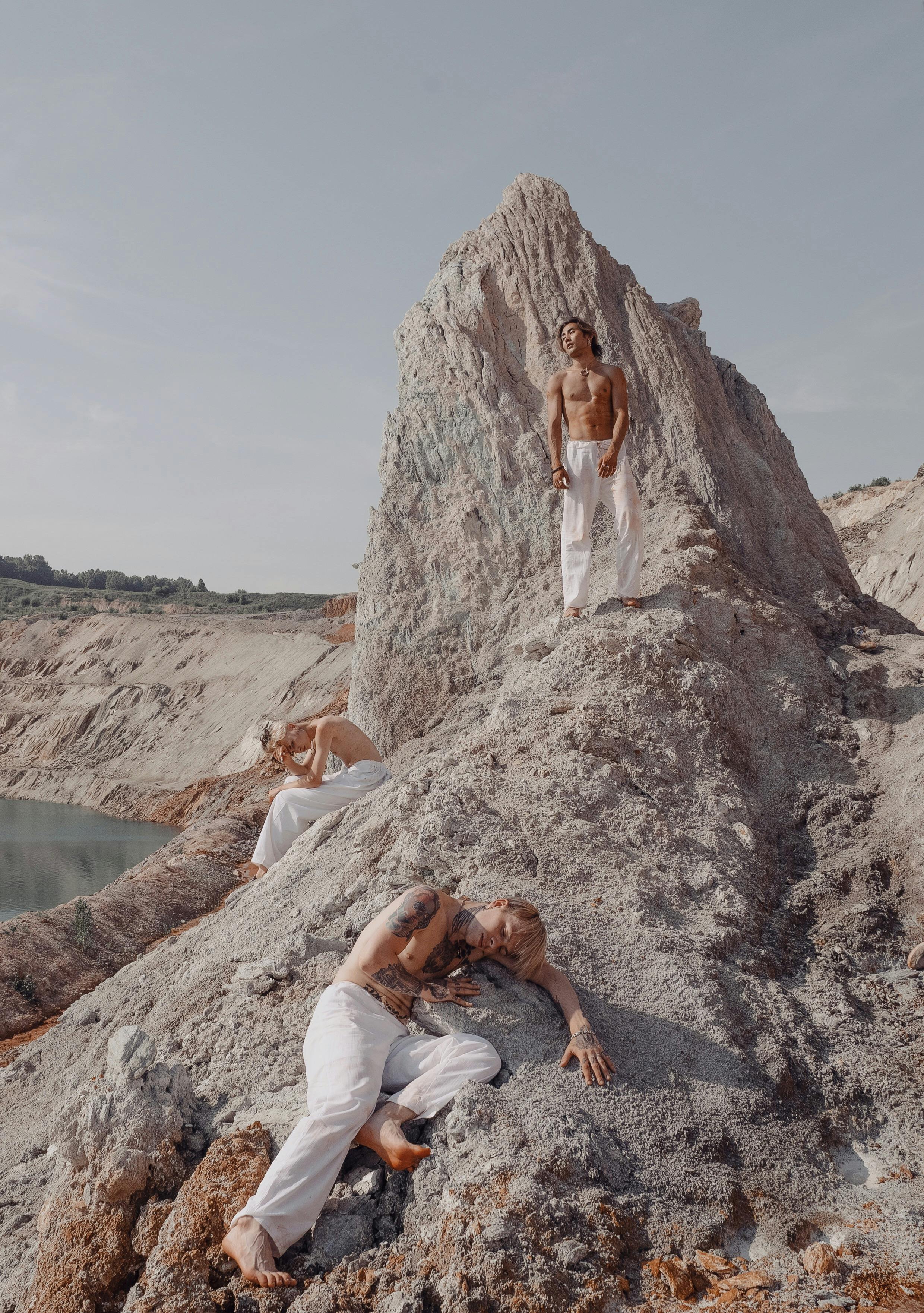 Three shirtless men with tattoos pose on a rocky outdoor terrain in bright daylight.