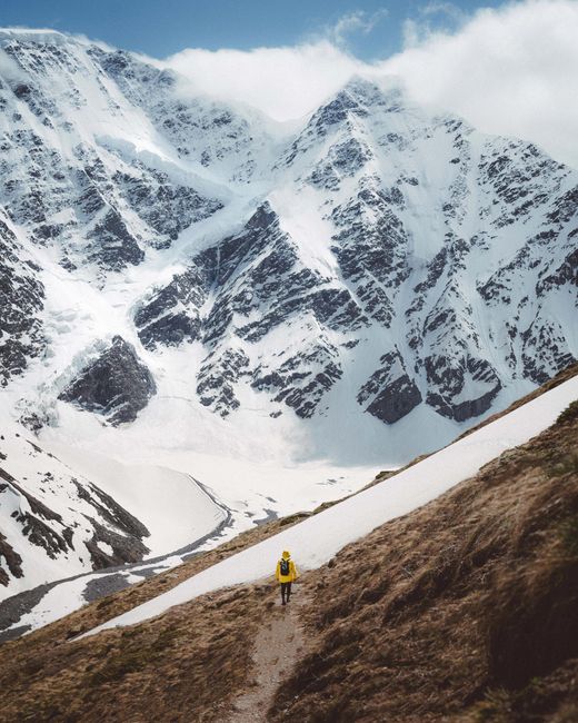 A lone trekker in bright gear traverses a snowy mountain path with a dramatic, cloud-covered peak.