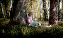 An Elderly Woman Sitting on a Green Grass while Leaning on a Tree Trunk