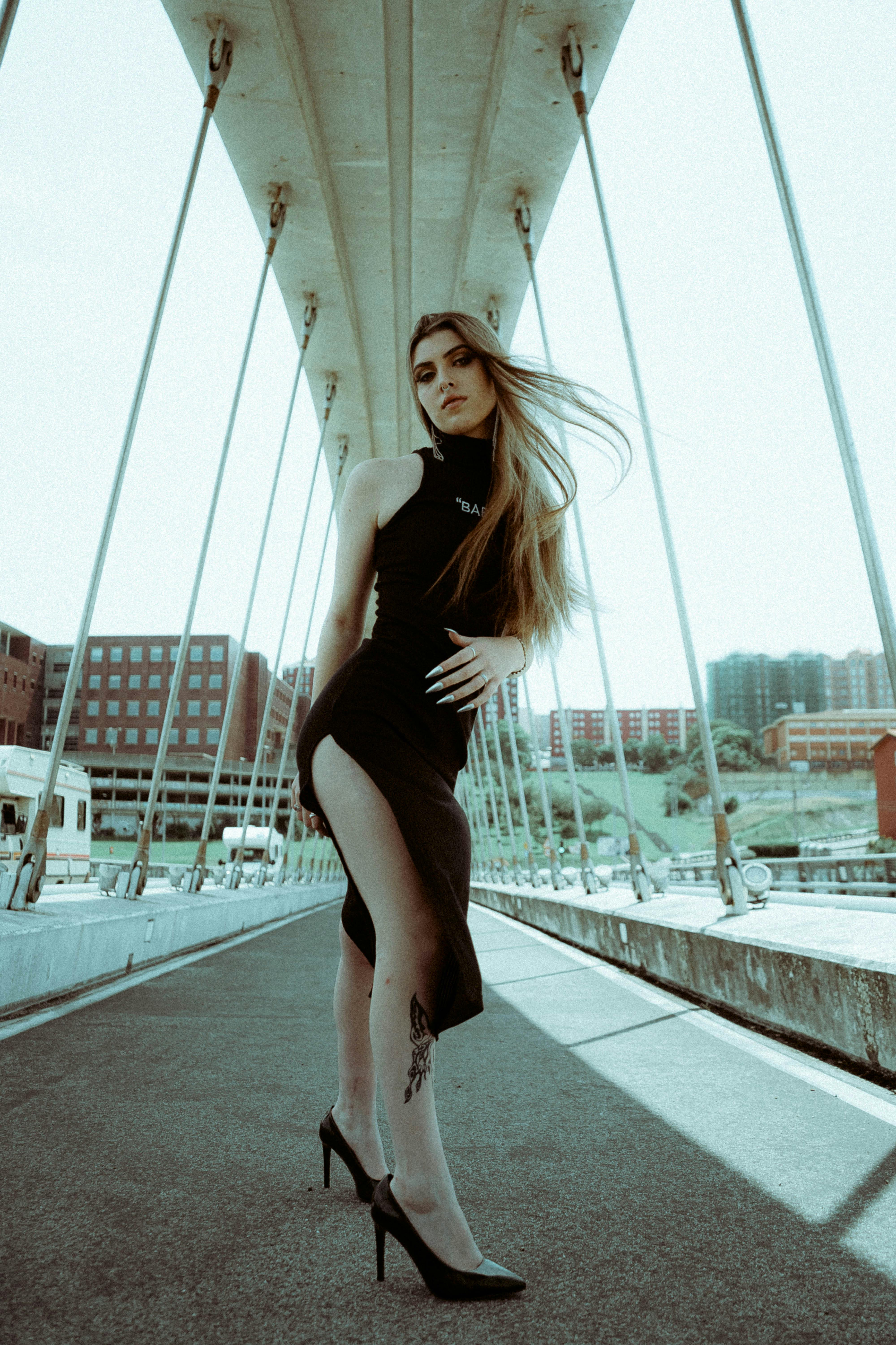 Stylish woman in a black dress poses on a modern urban bridge with striking architecture.