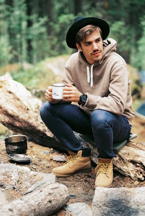 A Man in Denim Jeans Sitting on a Tree Log while Holding a Ceramic Mug