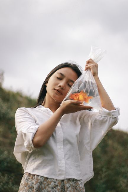 Woman holding a plastic bag with a goldfish against a natural outdoor background.