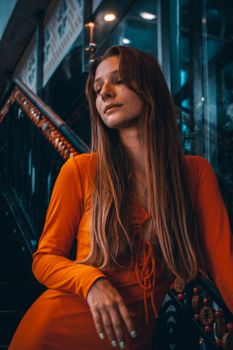 A stylish woman in an orange dress posing gracefully on a stairway indoors.