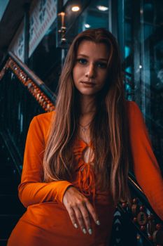 A beautiful woman in an orange dress poses gracefully on a dimly lit staircase, conveying elegance.