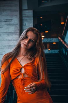 A woman in an orange top stands elegantly on a staircase, captured indoors.
