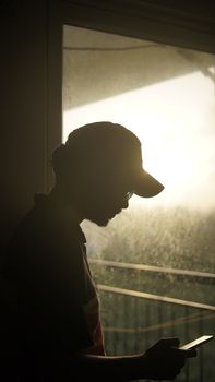 A silhouette of a man using a smartphone in front of a backlit window, capturing peaceful solitude.