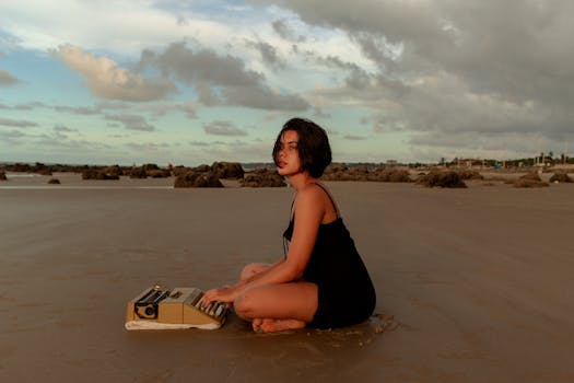 A woman in a black dress sits on a sandy Brazilian beach with a typewriter at dawn, surrounded by serene nature.