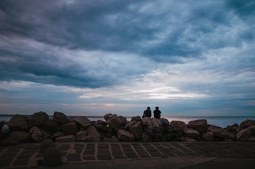 Two People Sitting on Coastal Rocks during Sunset