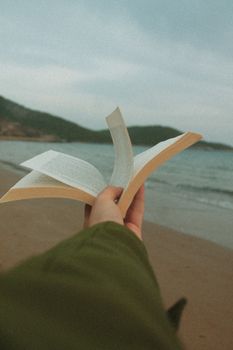 Close-up of an open book held by hand on a serene beach with calm sea.