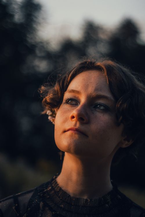 Free A woman gazes upwards thoughtfully in a moody outdoor setting, illuminated by warm twilight tones. Stock Photo
