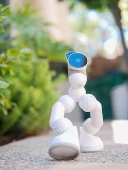 A small white robot toy on a concrete surface, surrounded by greenery.