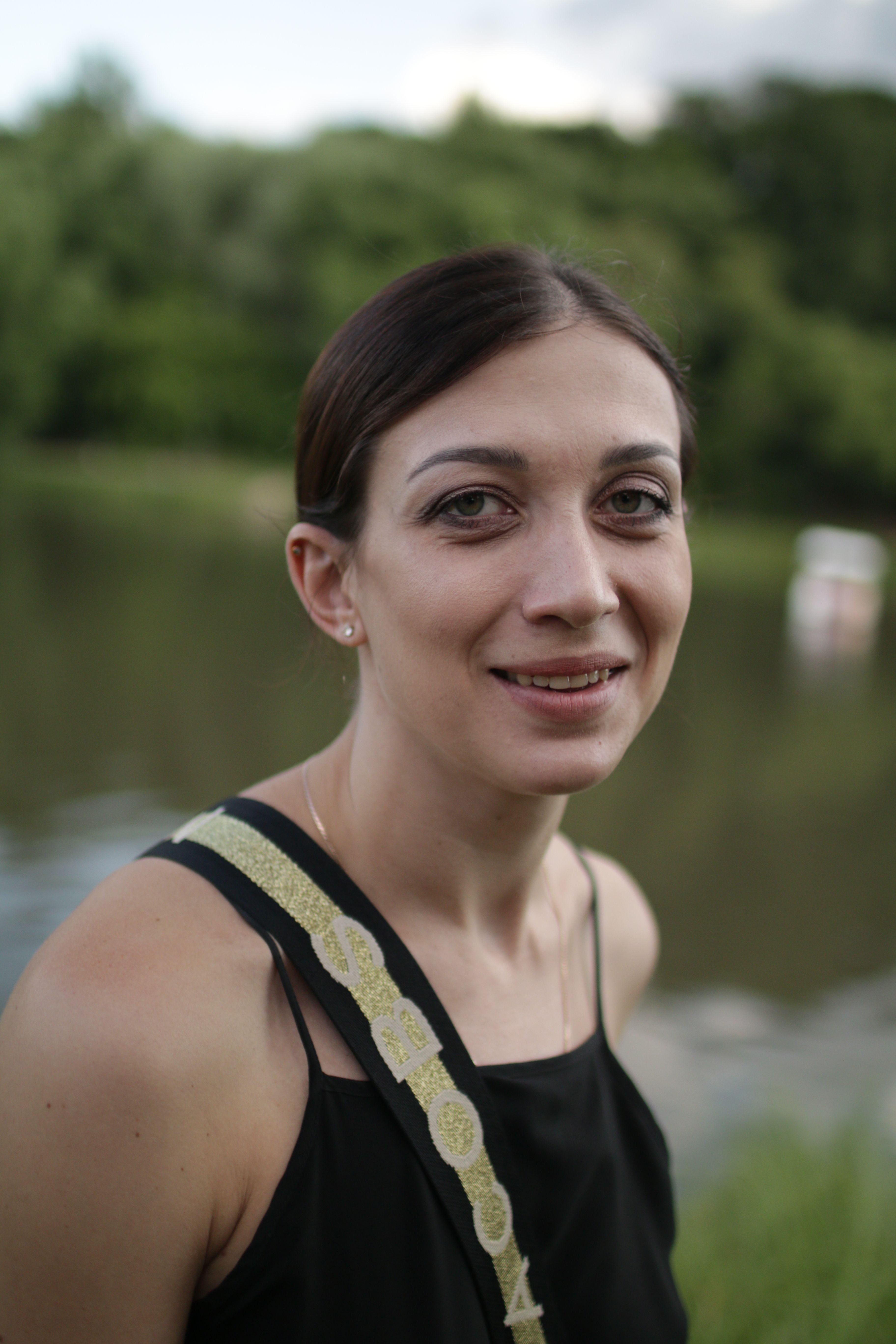 A smiling woman outdoors near a lake with a green forest background.