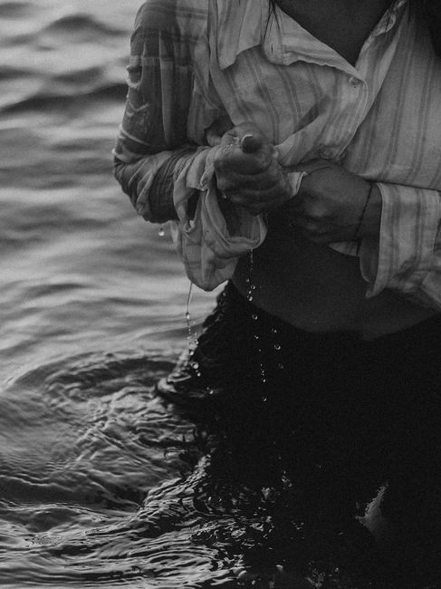 A moody black and white shot of a person wringing out wet clothing by the water's edge.