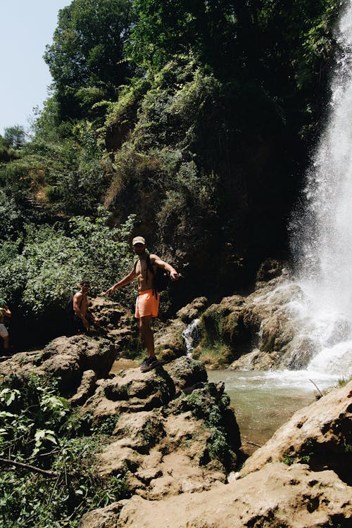 Man in Orange Shorts Standing on Rock Near Waterfalls