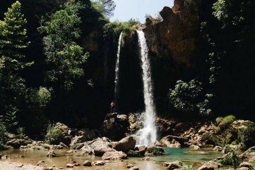 Majestic waterfall cascading into a clear stream surrounded by dense green forest under daylight.