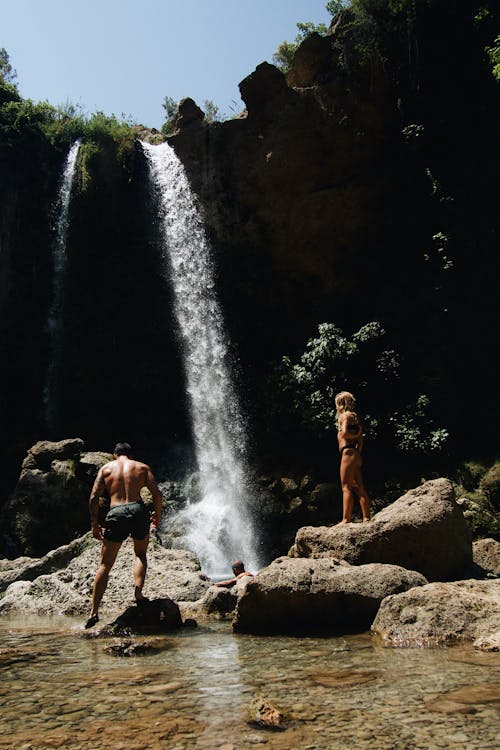 Woman in Black Bikini Standing on Brown Rock Near Waterfalls