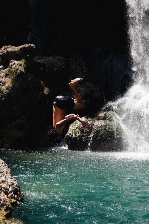 Man in Black Shorts Jumping Off the Cliff Beside Waterfall