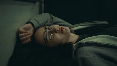 Free Woman in Eyeglasses Wearing Grey Hoodie and Lying with Hand Above Head Stock Photo