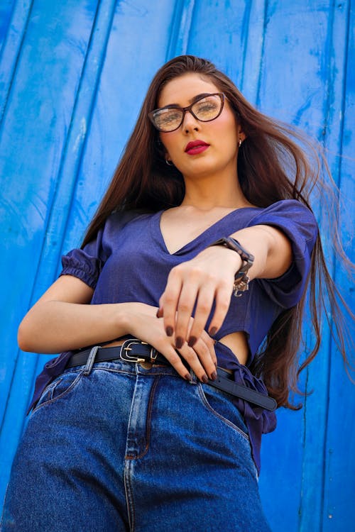 Woman in Framed Eyeglasses with Long Hair and Red Lips Standing Near Blue Wall with Extended Arm