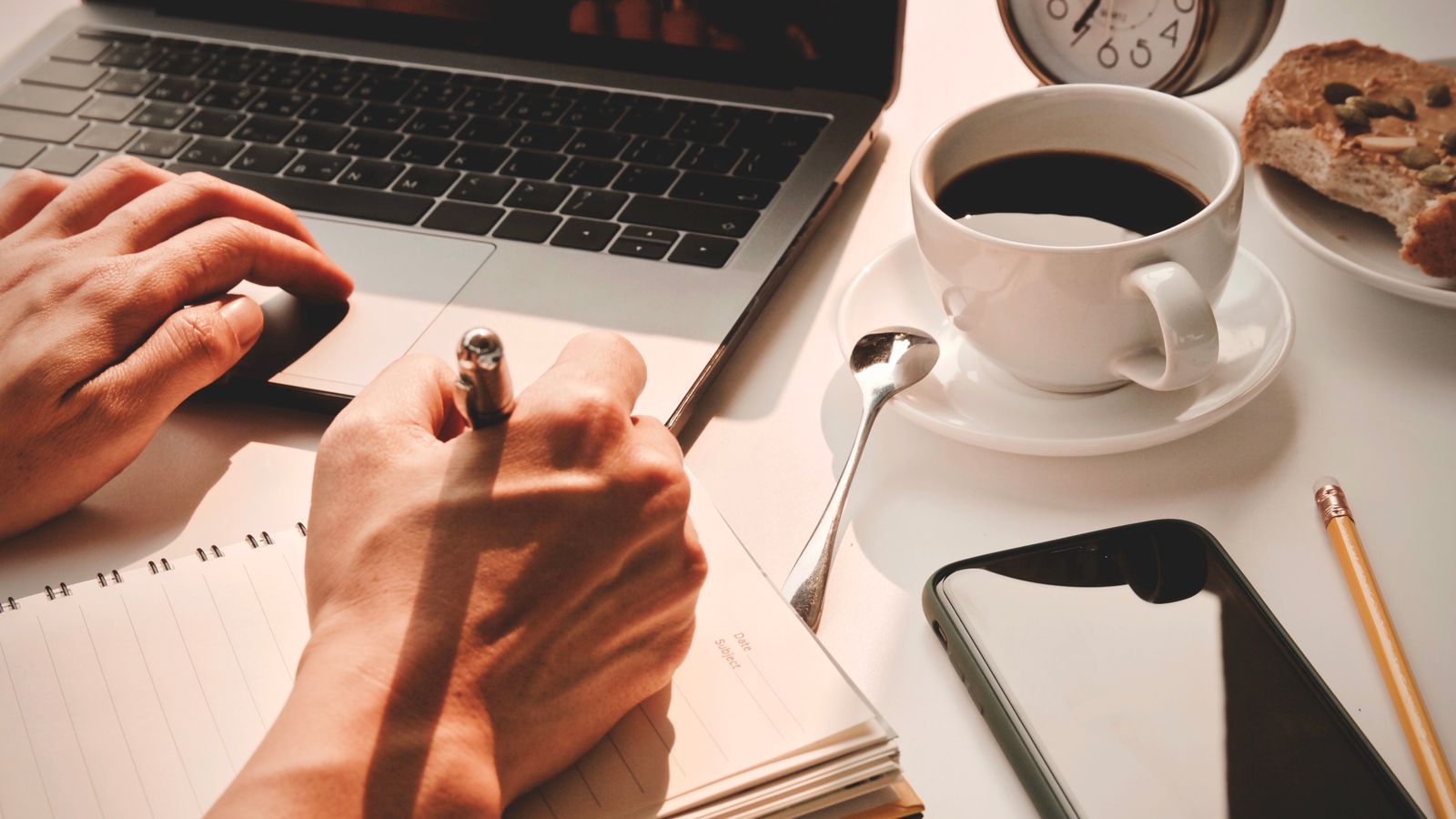 Close-up of hands writing in a notebook beside a laptop, smartphone, coffee and alarm clock