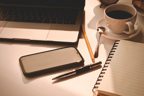 Free A warm workspace scene featuring a laptop, smartphone, coffee cup, and a notebook. Stock Photo