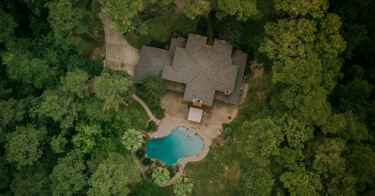 A scenic aerial shot of a house surrounded by lush forest and a swimming pool.