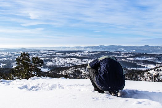 Person in winter attire kneels overlooking snowy landscape and distant hills under a clear sky.