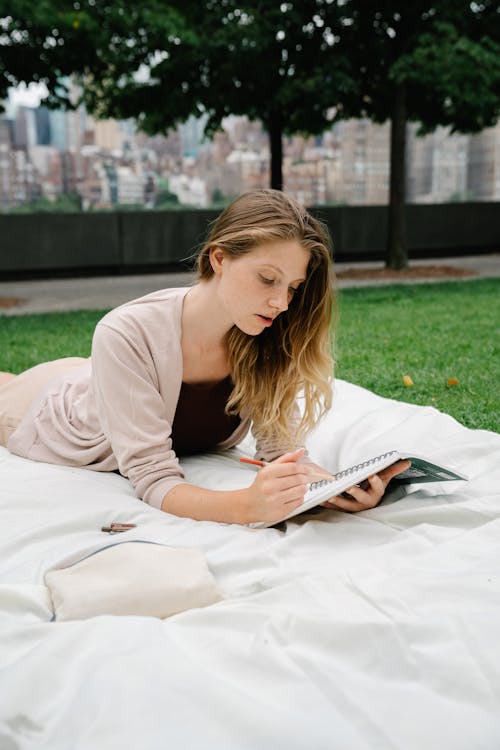 Woman Lying Down on a Blanket in a Park while Writing on a Notebook