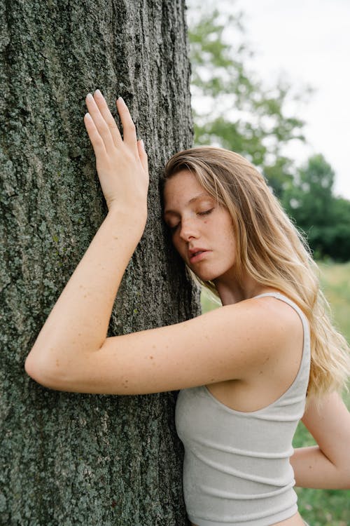 Woman in White Tank Top Leaning on Tree Trunk while Eyes are Closed