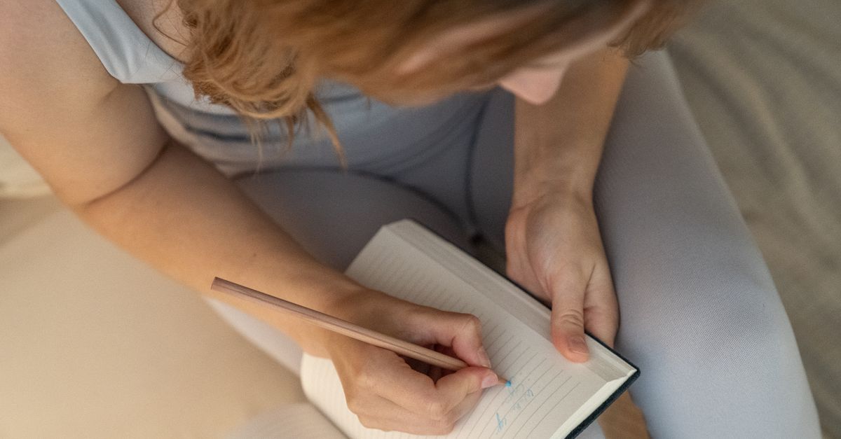 An adult woman writes in a notebook with a pencil, viewed from above.