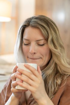 A blonde woman with eyes closed holding a steaming white mug indoors, enjoying a quiet moment.