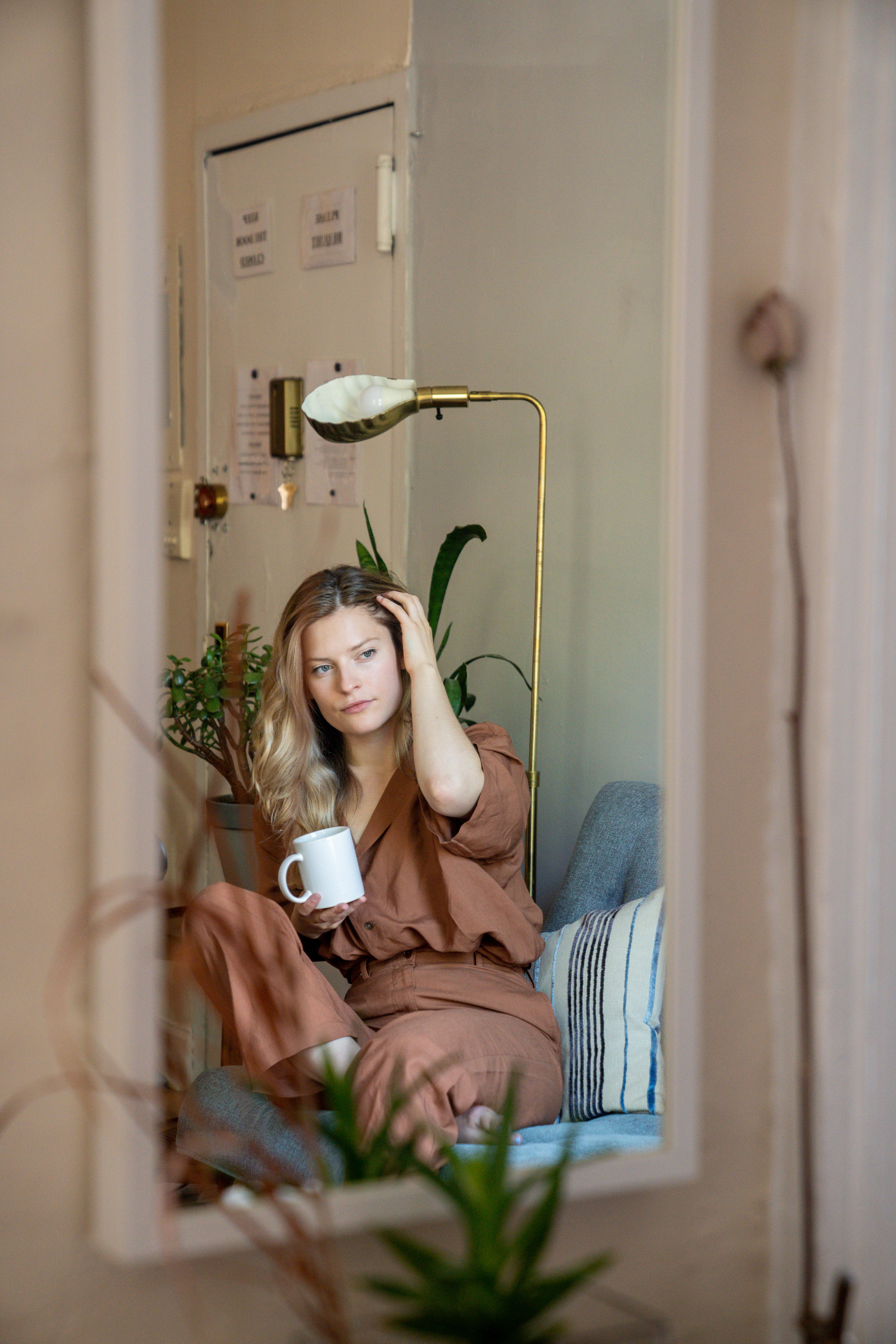 A woman in brown attire reflected in a mirror, relaxing indoors with a coffee mug.