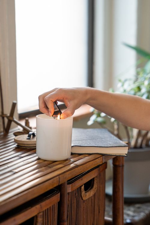 Free Hand lighting a white candle on bamboo table beside a cozy window. Stock Photo