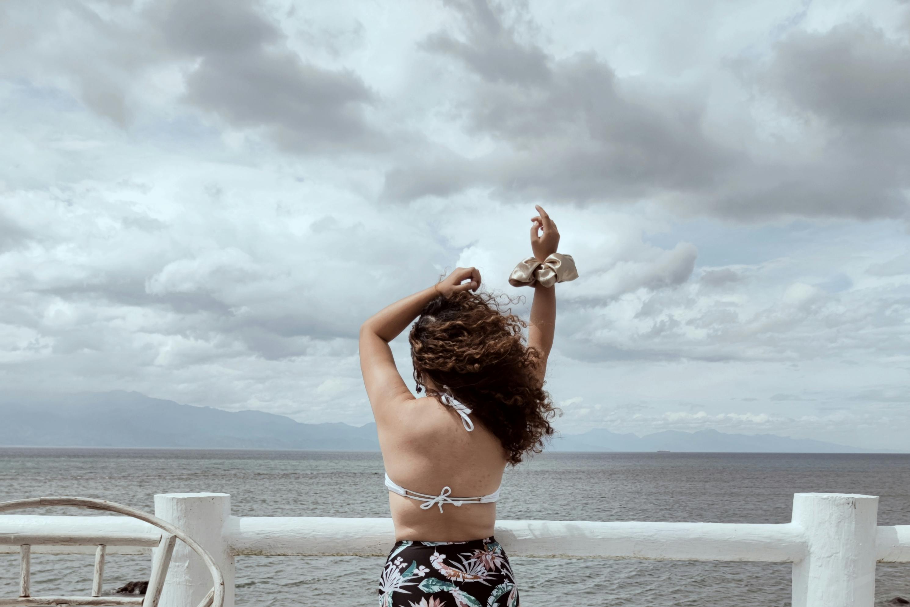 Rear view of a woman in swimsuit enjoying a serene ocean view with arms raised.