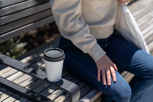 Free Close-up shot of a paper coffee cup on a park bench next to a person. Stock Photo