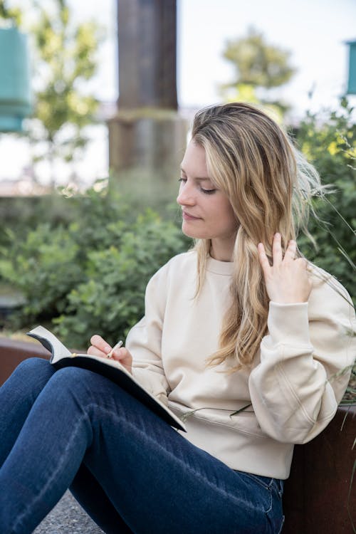 Free Woman in Sweater and Denim Pants Holding Her Hair While Writing on Notebook  Stock Photo