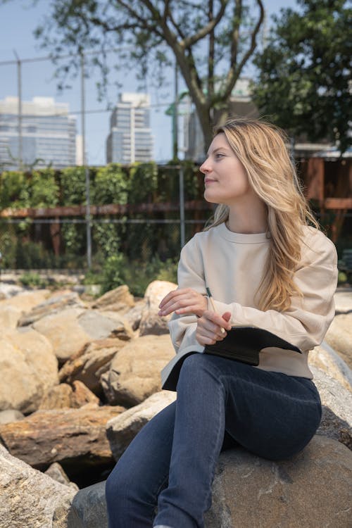 Free Woman Holding a Pen and Notebook While Sitting on Rock  Stock Photo