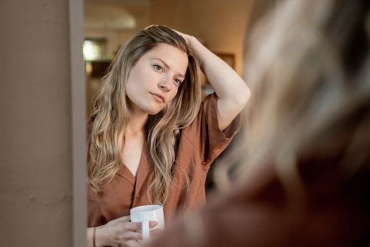 A woman gazes into a mirror, sipping coffee at home, with a serene expression and natural light accentuating her features.