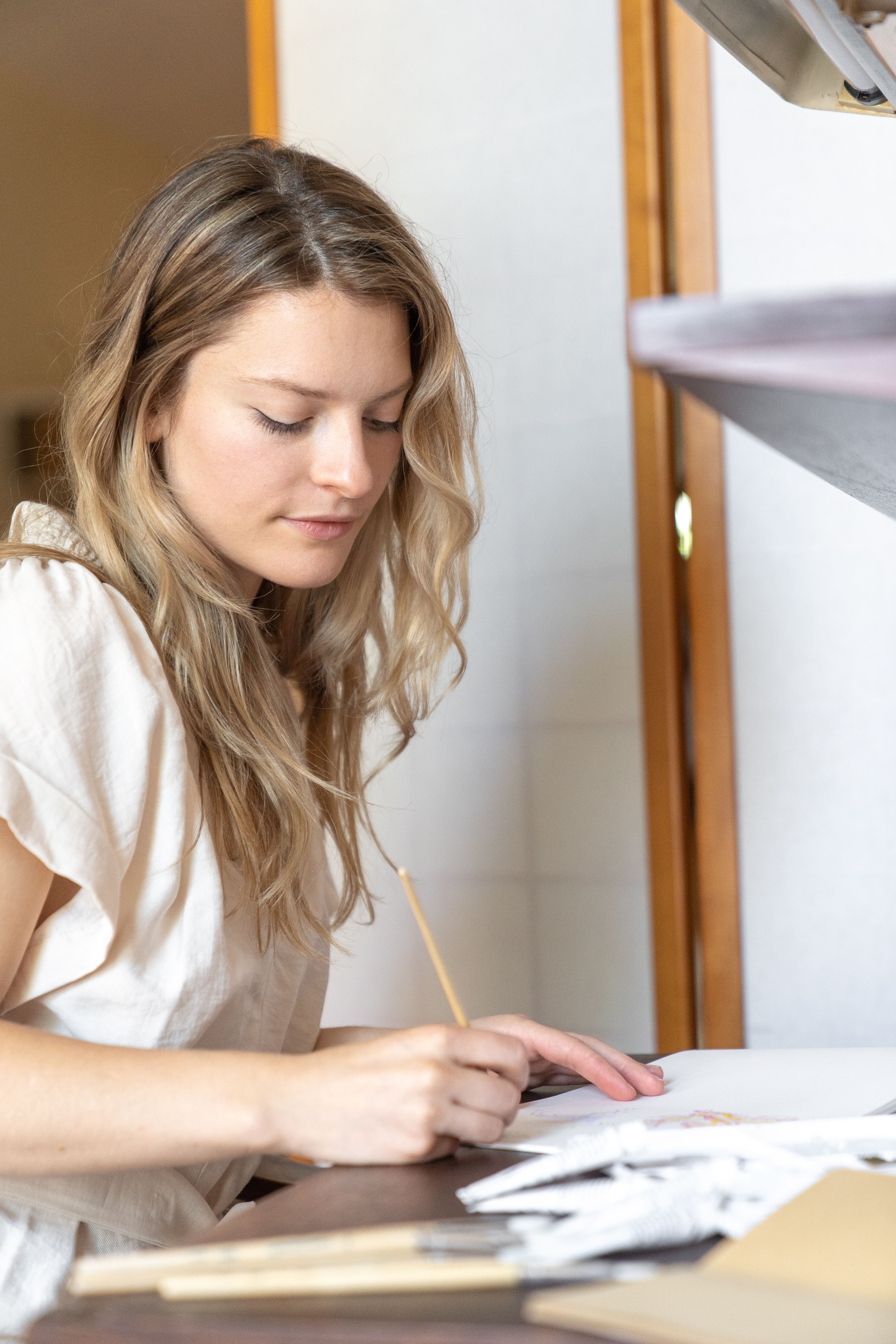 Young adult woman focused on writing in a notebook at a desk in a brightly lit indoor setting.