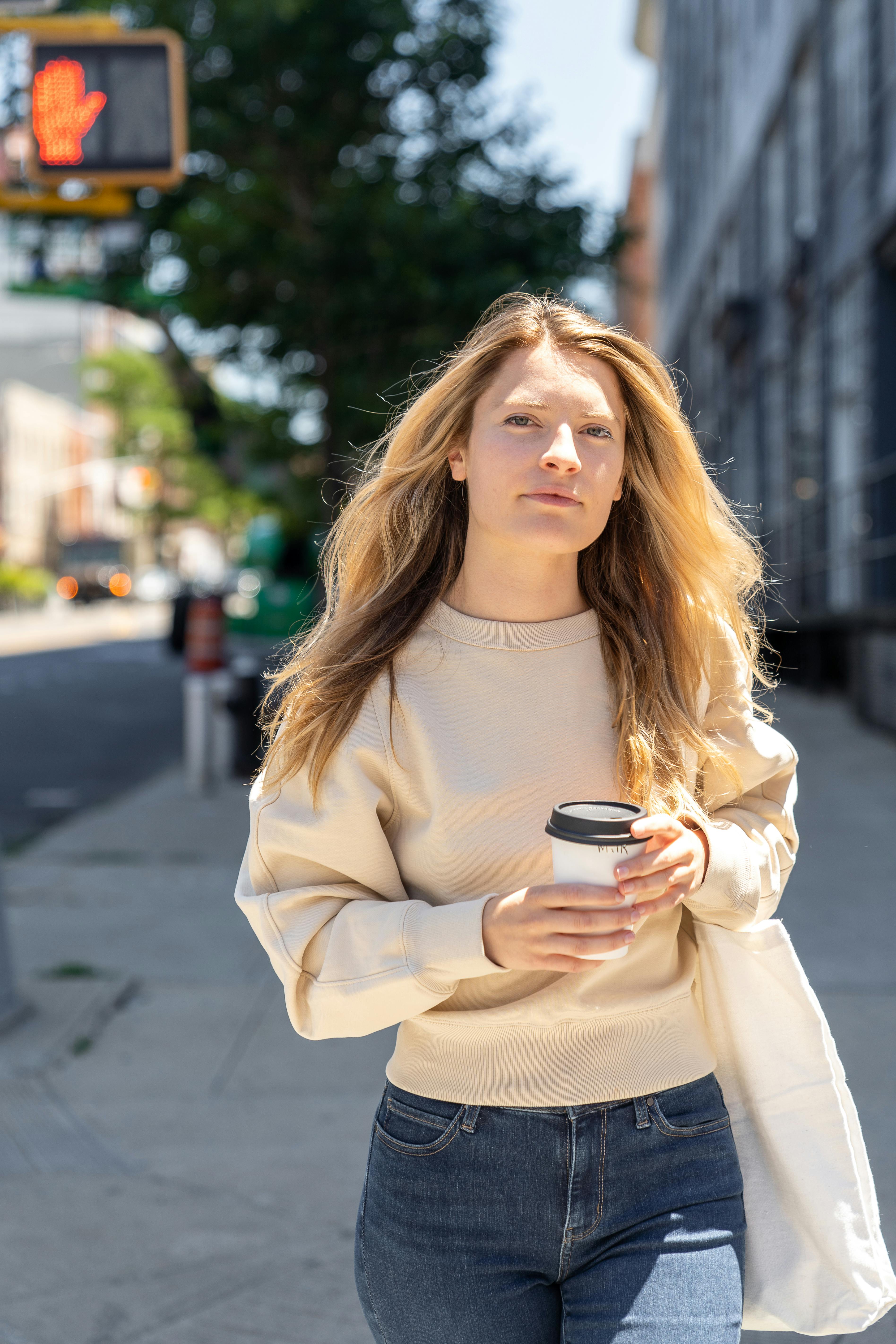 Casually dressed woman holding coffee walking on a city street during daytime.