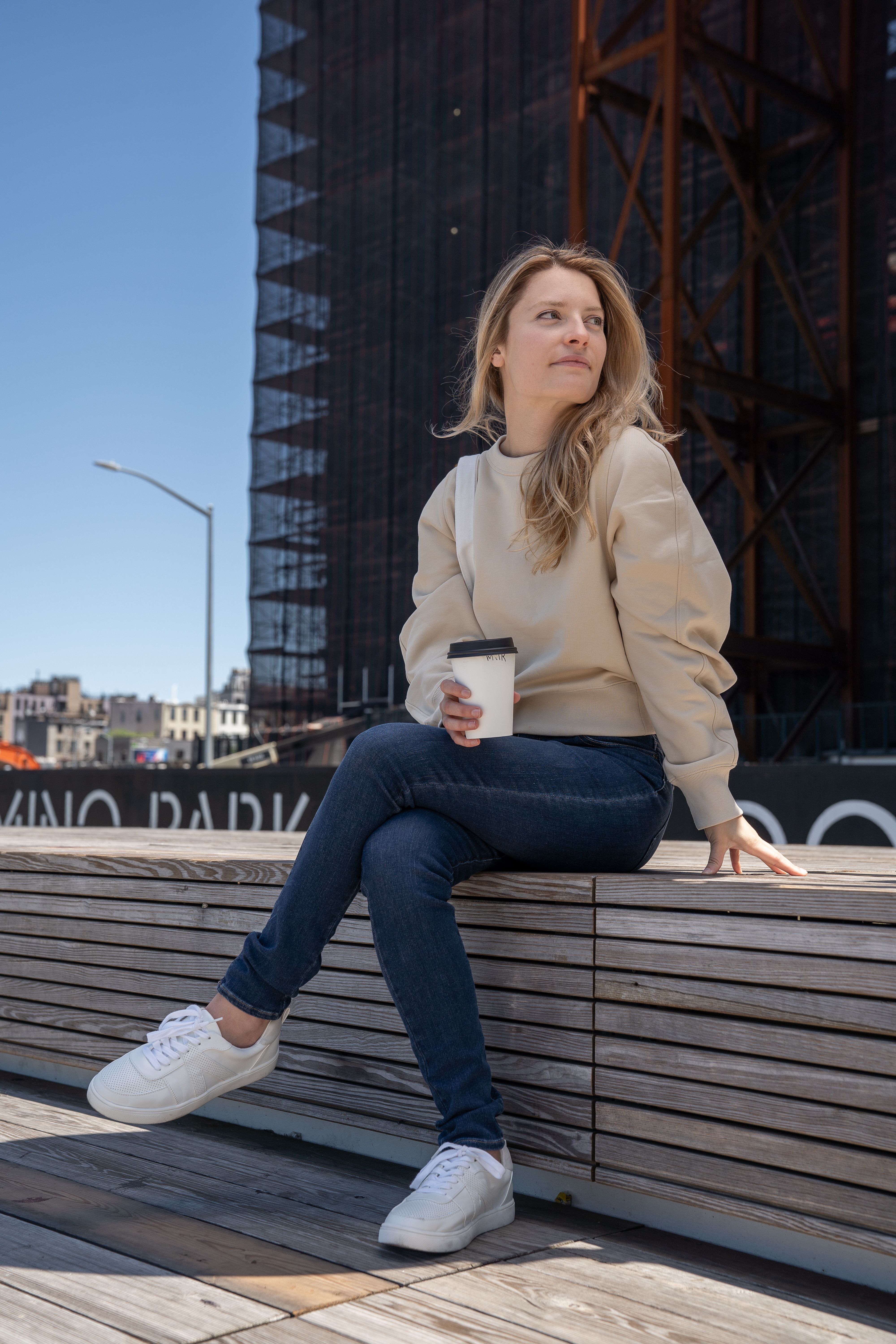 Casual young woman enjoying coffee outdoors in city setting with modern architecture background.