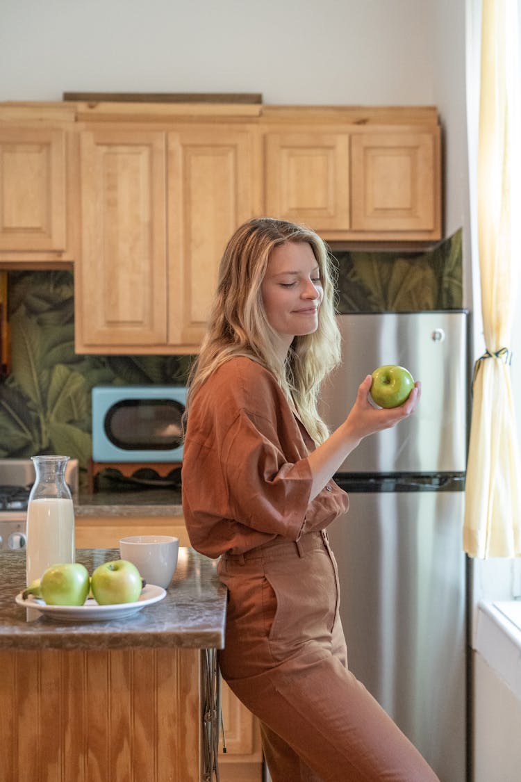 Smiling Woman Leaning Against The Kitchen Counter Eating An Apple 