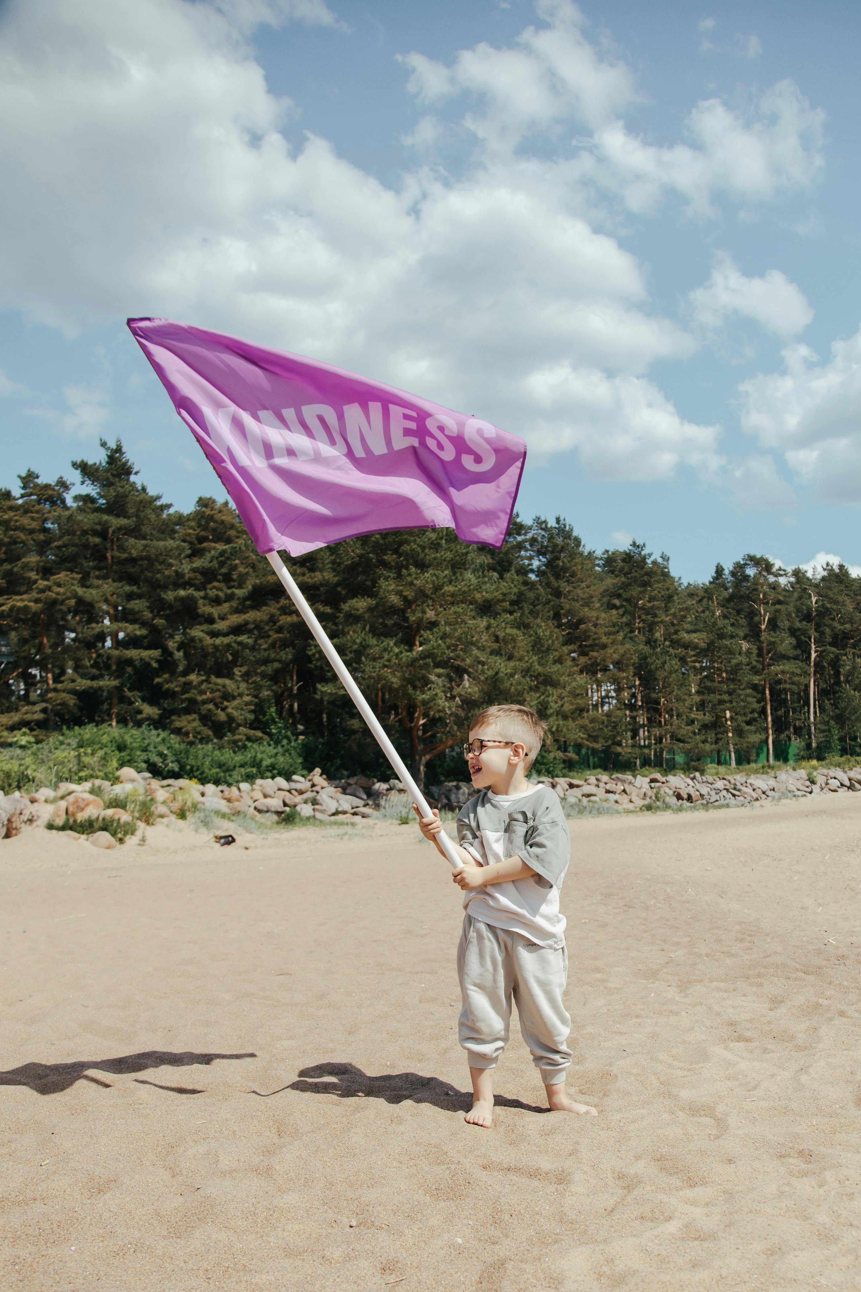 A young boy holds a large purple flag with 'Kindness' printed on it at a sandy beach under a clear sky.