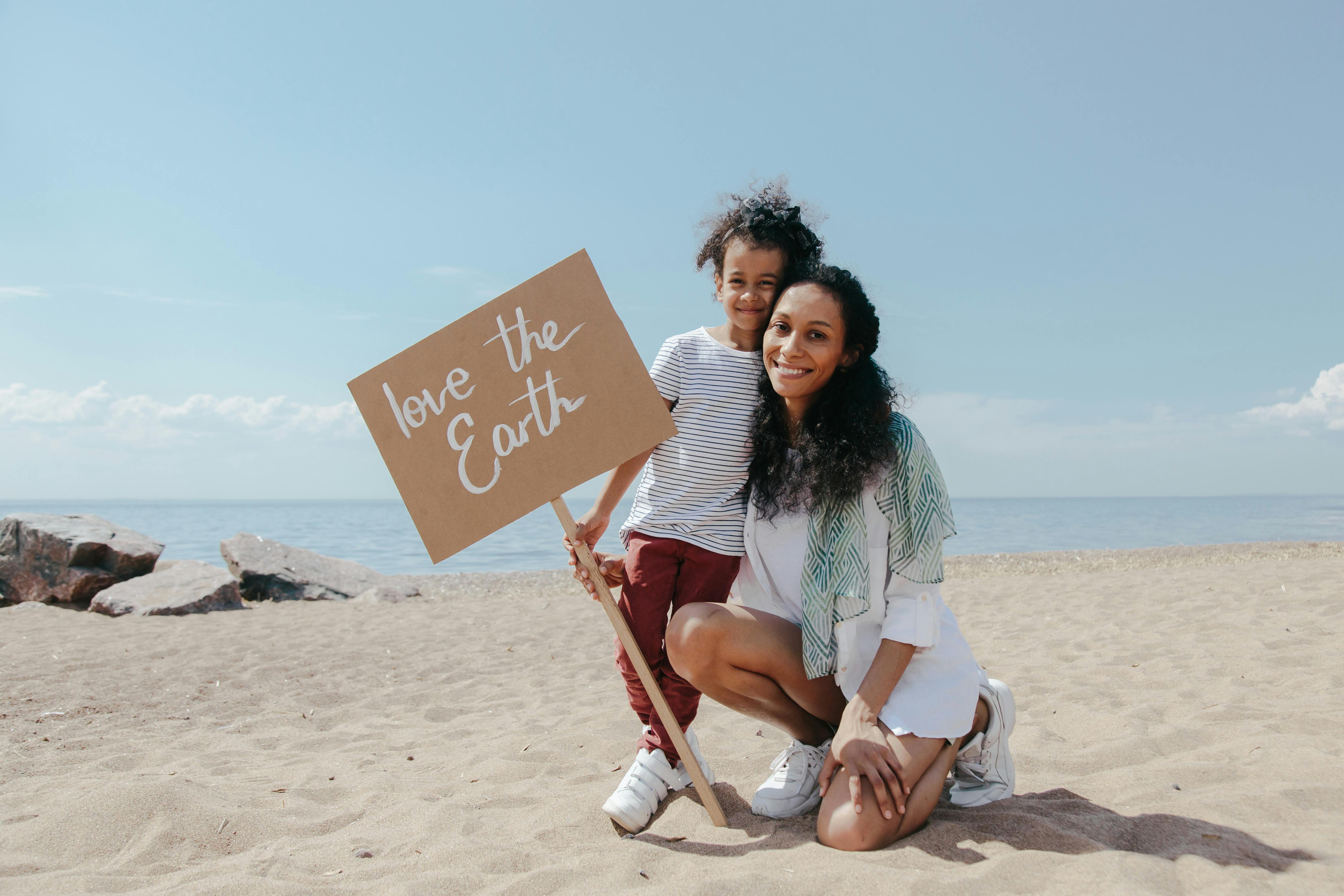 A mother and child holding a 'Love the Earth' sign on a sunny beach, promoting environmental awareness.
