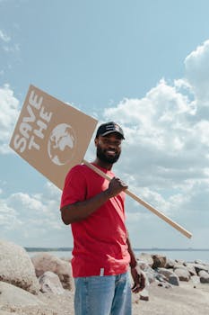 A man in a red shirt holding a 'Save The Planet' sign standing on a rocky beach under a blue sky.