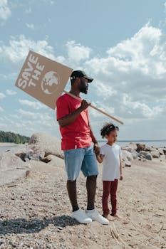 A man and child hold a Save the Planet sign on a rocky beach under a blue sky.
