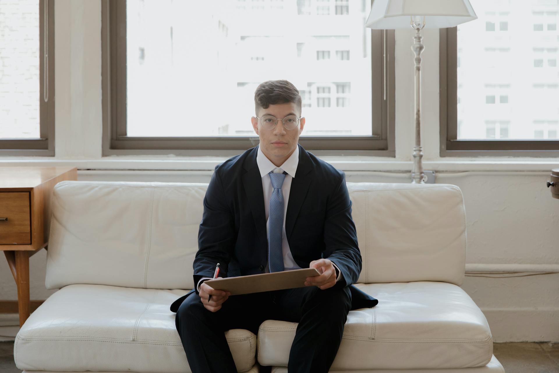 Young businessman in a suit sitting with a clipboard in a bright office setting. Ideal for corporate themes.